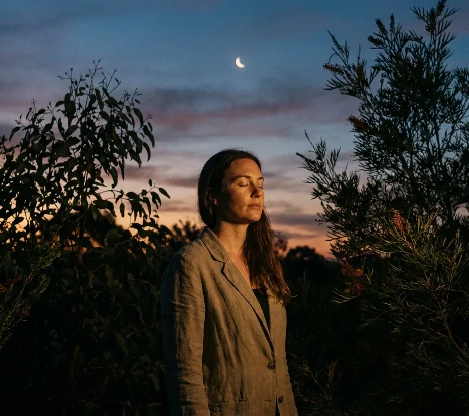 Woman standing among native shrubs at dusk with eyes closed, crescent moon above